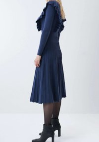 Robe marine côtelée avec encolure à volants, manches longues et jupe plissée évasée. Portée avec des bottines noires et des collants transparents.