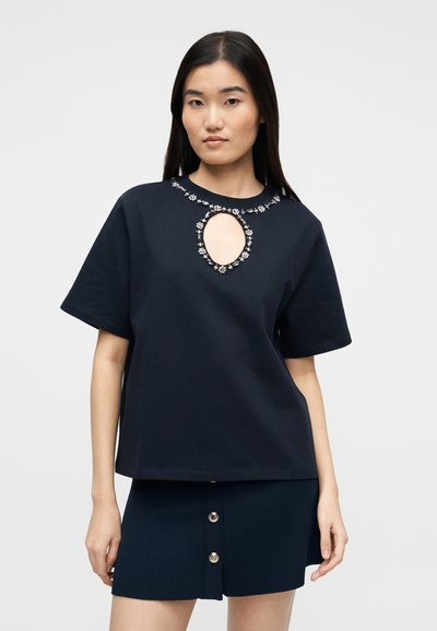 Νέα γυναίκα που φοράει μαύρο κοντομάνικο πάνω μέρος με λαιμόκοψη διακοσμημένη με κοσμήματα και άνοιγμα σε σχήμα κλειδαρότρυπας, σε συνδυασμό με μαύρη μίνι φούστα με κουμπιά.