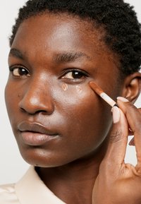 Correcteur liquide marron appliqué sous l'œil avec un pinceau, sur un modèle à la peau foncée. Texture de peau lisse, look de maquillage naturel léger.