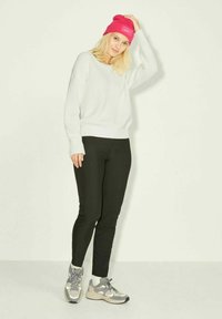Suéter branco com textura canelada, leggings pretas e sapatilhas cinzas com detalhes. Gorro rosa choque. Modelo posa contra um fundo simples.