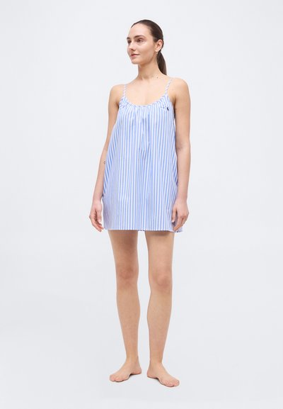 Γυναίκα που στέκεται ξυπόλυτη, φορώντας ένα χαλαρό φόρεμα με κάθετες μπλε και λευκές ρίγες, χωρίς μανίκια και με λεπτά τιραντάκια, πάνω σε απλό φόντο.
