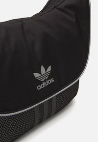 Borsa a tracolla nera in tessuto con una patta, caratterizzata da un logo Adidas argentato e un motivo a tre strisce orizzontali nella parte inferiore. Superficie texture.