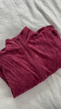 Veste à capuche zippée bordeaux, fabriquée en tissu texturé avec un motif côtelé, présentant un petit logo sur le côté gauche et une finition douce.