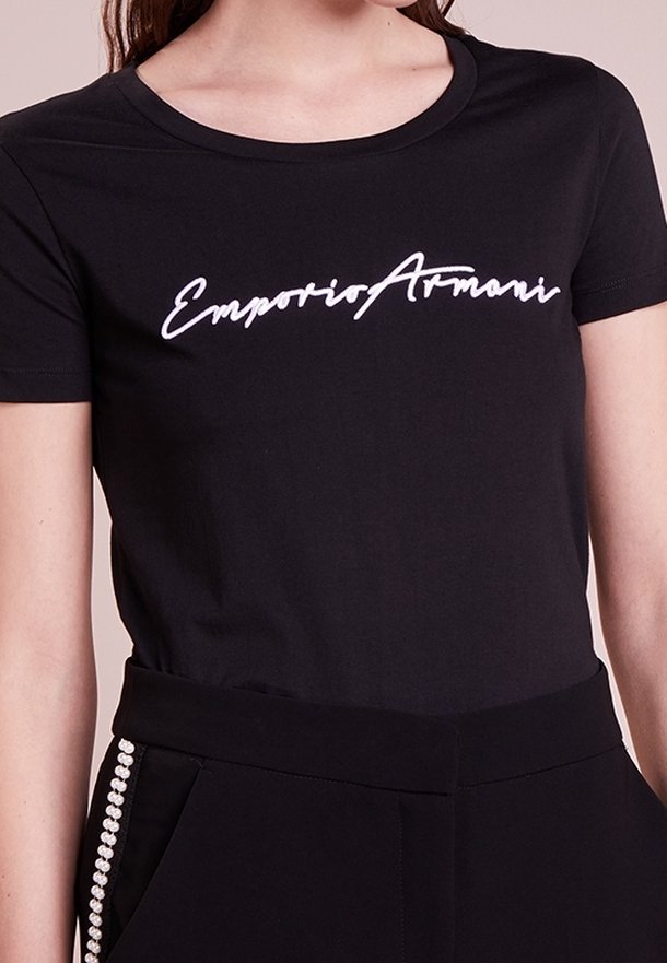 Camiseta negra de manga corta ajustada con el logotipo en blanco "Emporio Armani" usada con pantalones negros que presentan detalles de perlas en el costado.