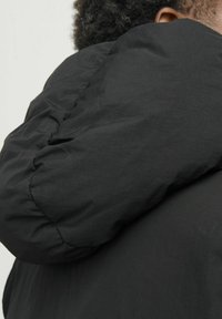 Schwarze gesteppte Jacke mit hohem, rundem Kragen. Das Material wirkt glatt mit leichtem Glanz und zeigt weiche, geraffte Texturen.