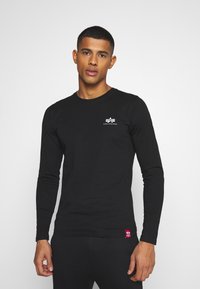 Langarm-T-Shirt in Schwarz aus weichem Baumwollstoff, mit einem kleinen weißen Logo auf der Brust und einem roten Stoffetikett am Saum.