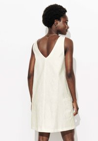 Robe sans manches blanche avec un design en V dans le dos, motif texturé et poche latérale, avec un ourlet au niveau des genoux.