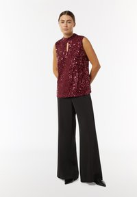 Bordeaux farbener Paillettenoberteil mit hohem Kragen und Schlüssellochdetail, kombiniert mit schwarzen weit geschnittenen Hosen und spitzen schwarzen Schuhen. Seidige Textur.