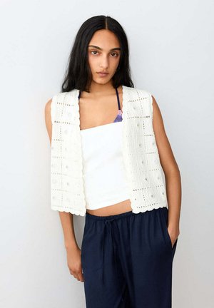 Femme aux longs cheveux foncés porte un gilet blanc en crochet sur un top tube blanc, accompagné d’un pantalon ample bleu marine, debout devant un fond uni.