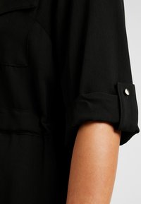 Zwarte shirt met opgerolde mouwen en drukknoop, gemaakt van een gladde stof. Inclusief een borstzak en een gestructureerd silhouet.