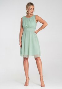 Miętowa sukienka bez rękawów z dopasowanym korpusem, marszczeniami w talii oraz przezroczystą, warstwową spódnicą. Gładka faktura materiału.