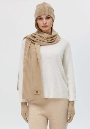 Béret en tricot beige et écharpe côtelée avec une étiquette, associés à un pull blanc pelucheux et un pantalon beige assorti, mettant en valeur une texture douce.