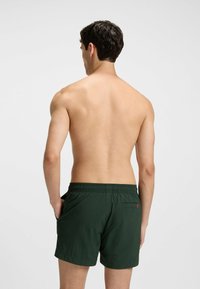 Costumi da bagno verdi con cintura elastica, tasche laterali e posteriori, e una texture liscia, mostrati da dietro su uno sfondo chiaro.