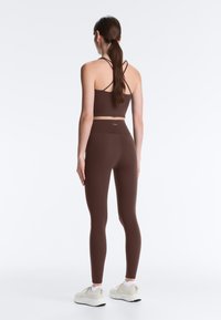 Woman with long brown hair in a ponytail wearing brown crisscross strap crop top and matching leggings, standing facing away on a white background.