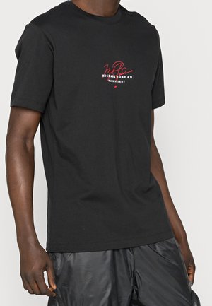 Chemise noire à manches courtes avec texte rouge et blanc "Michael Jordan Take Flight" et logo signature, portée par une personne à la peau foncée.