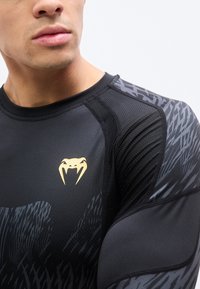 Κοντινό πλάνο ενός άνδρα που φοράει μαύρο αθλητικό πουκάμισο με δίχτυ και μανίκια με σχέδιο, με χρυσλό λογότυπο φιδιού με δόντια στο στήθος.