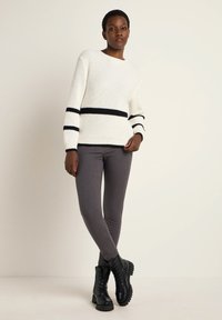 Weißer Strickpullover mit schwarzen Streifen, kombiniert mit grauen, figurbetonten Hosen. Schwarze Schnürstiefel haben eine strukturierte Sohle und ein robustes Design.