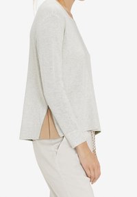 Maglione grigio chiaro a maniche lunghe con scollatura rotonda, fessure laterali e una vestibilità leggermente drappeggiata; realizzato in un materiale morbido e testurizzato.