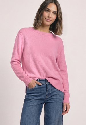 Femme portant un pull en maille rose et un jean bleu, debout avec une main dans la poche et un léger sourire sur un fond uni.