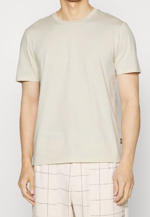 Männliche Person trägt ein hellbeiges T-Shirt mit kurzen Ärmeln und Rundhalsausschnitt sowie hellfarbene Shorts mit schwarzem Gittermuster, steht mit entspannt an den Seiten hängenden Armen.