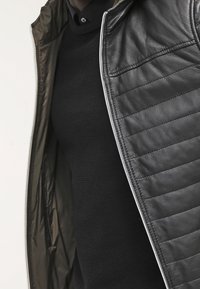 Giacca di pelle nera con texture trapuntata, caratterizzata da una chiusura con zip e rivestimento verde oliva. Indossata sopra un maglione nero in maglia.