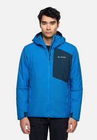 Blauwe geïsoleerde jas met een donkerblauwe borstzak, ritssluiting en een capuchon. Beschikt over textuurstof en een merkembleem op de zak.