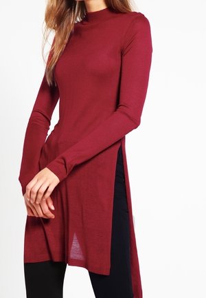 Kobieta ubrana w bordowy, długi sweter-tunikę z długim rękawem i wysokim kołnierzem, z bocznymi rozcięciami, w połączeniu z czarnymi spodniami, na białym tle.