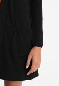 Svart kofta med strukturerad stickning, långa ärmar, ribbade manschetter och två framfickor, med en slät yta.