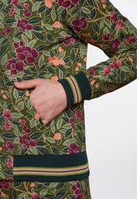 Sudadera con estampado floral en verde, con flores moradas y naranjas, puños acanalados con rayas en verde, morado y amarillo, y bolsillo lateral.