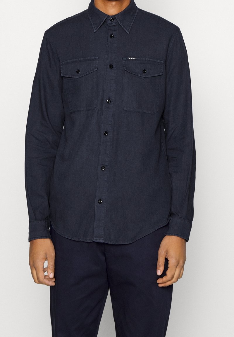 Tmavě modrá košile s dlouhým rukávem z bavlny, zapínáním na knoflíky vpředu, dvěma náprsními kapsami a černými knoflíky. Hladká textura.