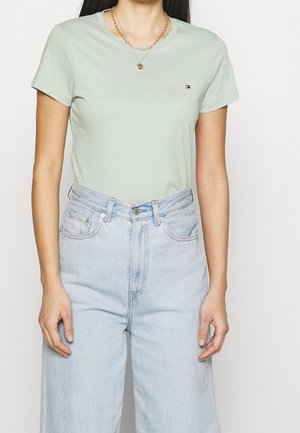 Vrouw draagt een lichtgroen T-shirt met korte mouwen en lichtblauwe hooggetailleerde spijkerbroek met twee gouden kettingen.
