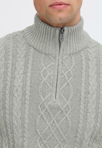 Light gray cable-knit sweater with a high ribbed collar and a zippered neck detail. Features thick and thin braided patterns.