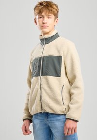 Giacca in fleece color crema con un pannello verde scuro e chiusura a zip. Presenta tasche laterali e colletto alto. Tessuto morbido e testurizzato.