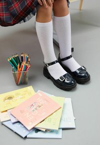 Zapatos de charol negros con una suela gruesa y hebilla, combinados con calcetas blancas hasta la rodilla; rodeados de cuadernos de dibujo coloridos y lápices.