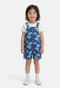 Jeune enfant debout, portant un t-shirt blanc, une salopette courte en denim avec un motif floral blanc, des chaussettes blanches et des baskets blanches, souriant légèrement.