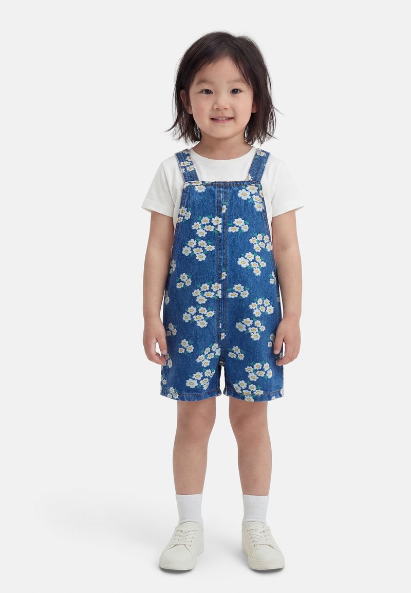 Jeune enfant debout, portant un t-shirt blanc, une salopette courte en denim avec un motif floral blanc, des chaussettes blanches et des baskets blanches, souriant légèrement.