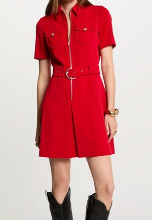 Robe zippée rouge avec des manches courtes, deux poches poitrine, ceinture avec boucle circulaire, jupe plissée, portée avec des bottes de cowboy noires.