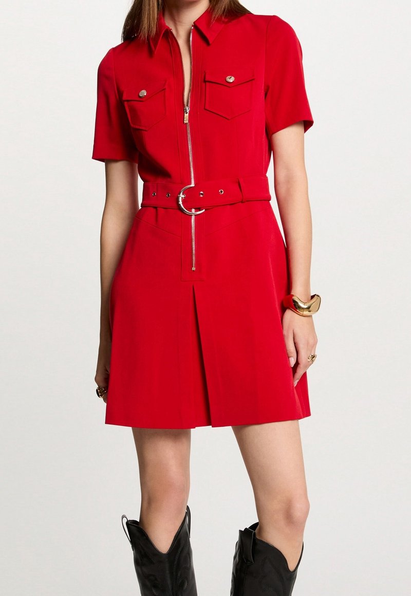 Rode zip-up jurk met korte mouwen, twee borstzakken, riem met ronde gesp, geplooide rok, gedragen met zwarte cowboylaarzen.