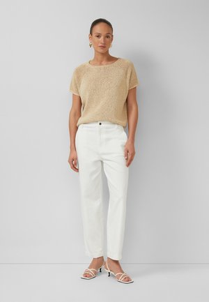 Donna in piedi contro una parete liscia che indossa un top a maniche corte beige in maglia, pantaloni bianchi a vita alta, sandali bianchi con cinturino e orecchini dorati.