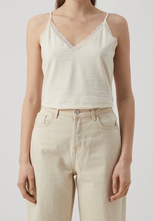 Kvinde iført en hvid ærmeløs top med blonderkant og beige højtaljede jeans, stående mod en ensfarvet baggrund.