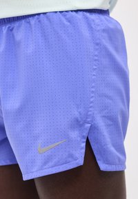 Κοντινό πλάνο ενός ατόμου που φοράει μοβ σορτς Nike με μικρές οπές και ανακλαστικό λογότυπο Swoosh στο αριστερό πόδι.