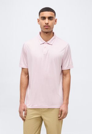 Mand med kort hår iført en lyserød polo og beige bukser, der står foran en ensfarvet lys baggrund.