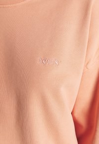Sudadera de un suave color durazno, confeccionada con un tejido blando, que presenta un pequeño logo bordado de Levi's en color rosa en la parte izquierda del pecho.