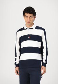 Mørkeblå og hvitstripet langermet polo skjorte med krage og et lite logo-lapp på brystet. Ribbestrikkede ermer og kant.