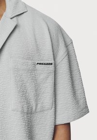 Hellgraues Kurzarmhemd mit strukturierter Stoffqualität, einem Kragen und einer Brusttasche mit dem schwarzen "PEGADOR"-Logo.