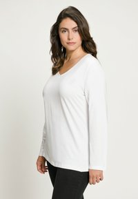 Bílý top s dlouhým rukávem a výstřihem do V, vyrobený z hladkého a měkkého materiálu. Má volný střih a rovný lem, kombinovaný s tmavými džíny.