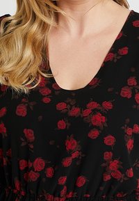 Svart klänning med en djup V-ringning som har ett blommönster av röda rosor. Tyget verkar mjukt med ett gathered midja.