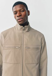 Beige lynlåsjakke lavet af struktureret materiale, der har to brystlommer med lynlås og et højt kravedesign.