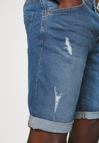 Shorts en denim bleu avec un design déchiré, présentant des patchs effilochés et des ourlets roulés. Le tissu a une texture lisse et un léger dégradé.