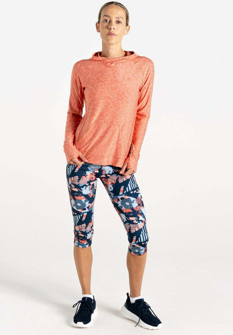 Langärmliges, korallenfarbenes, strukturiertes Oberteil mit einem Wickelausschnitt, kombiniert mit knielangen, gemusterten Leggings in Marineblau, Rosa und weißen geometrischen Designs.